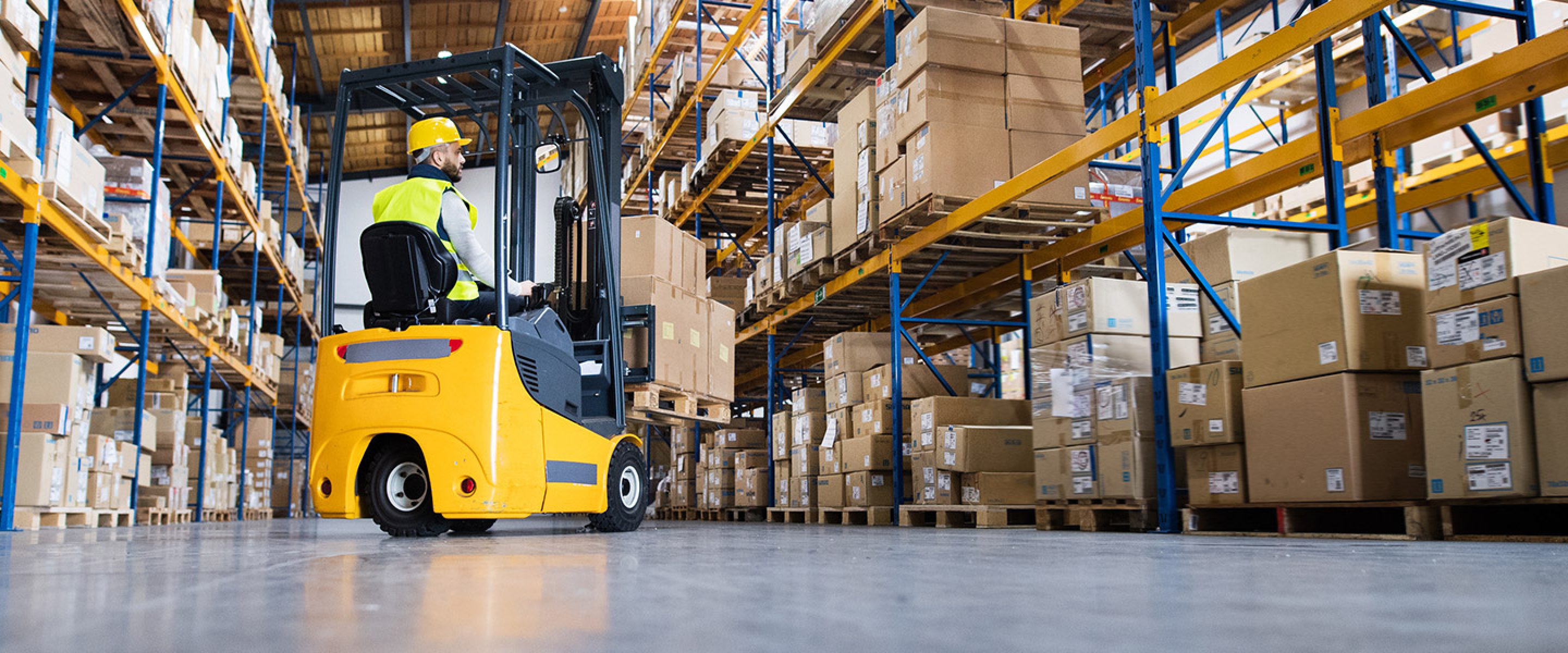 Male factory worker drives fork lift through aisles of shelved boxes